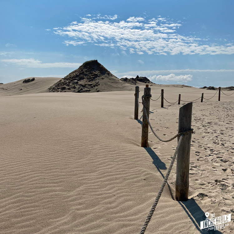 Am rechten Rand zieht sich eine Absperrung mit Holzpfosten und einem Seil bis zum Horizont, links viel Sand und ein Hügel aus Sand