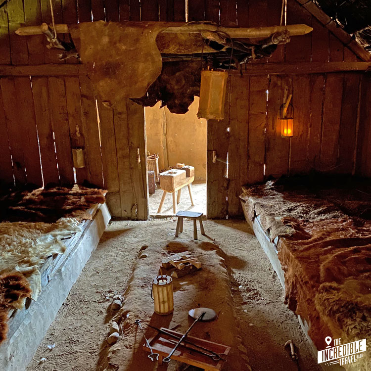 Ansicht eines Innenraums in Haithabu mit Schlafstätten links und rechts, in der Mitte ein flacher Tisch, über dem ein paar Felle aufgehängt sind