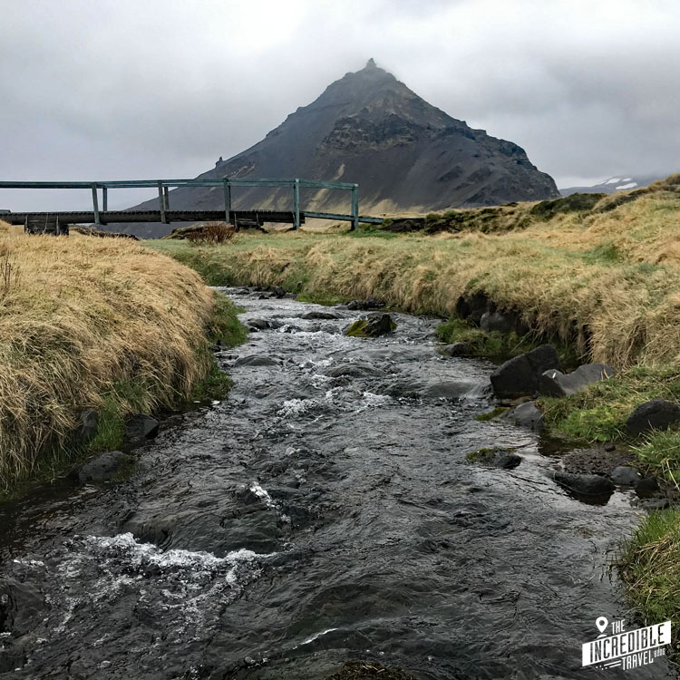 Fluss mit einer Holzbrücke im Vordergrund, im Hintergrund ein vulkanischer Berg