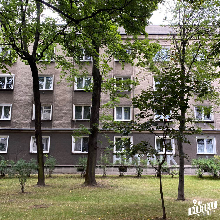 Blick durch einen Grünstreifen mit Bäumen auf ein graues Wohngebäude mit der Graffiti-Aufschrift "Hutnik"