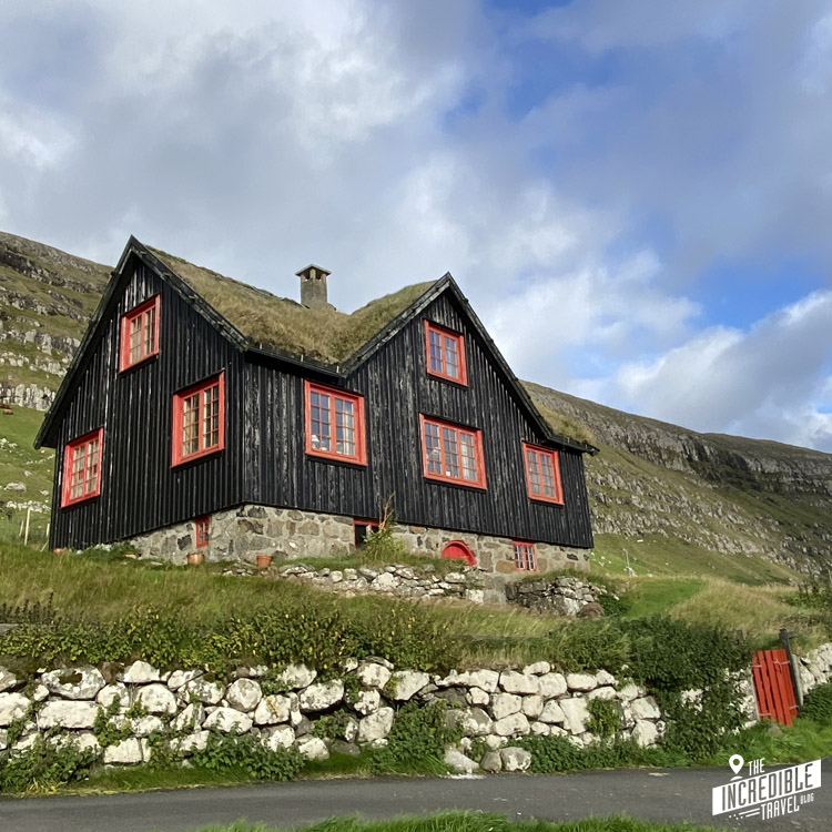 Schwarzes Holzhaus mit Natursteinfundament und roten Fensterrahmen und Grasdach