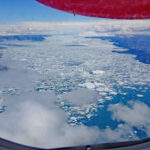 Blick aus dem Flugzeugfenster auf einen von Eischollen und Eisbergen fast vollständig bedeckten Fjord in Grönland