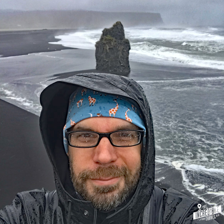 Selfie im Regenmantel, im Hintergrund ein schwarzer Strand mit einer vulkanischen Felsnadel