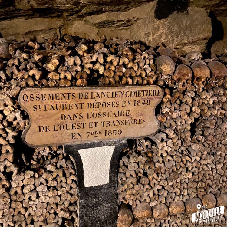 Hinweisschild auf den Herkunftsfriedhof vor einer Wand aus menschlichen Knochen und Schädeln