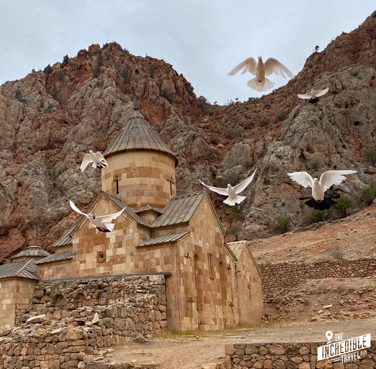 Klosterkirche vor einer Felswand, Tauben fliegen auf den Betrachter zu