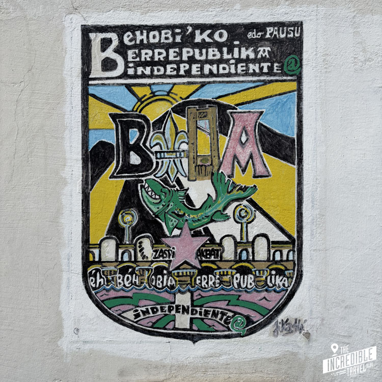 Street art mural with the text Berrepublika Independiente and B.I.A. in stylized letters. It features abstract designs with a star, green dragon, and landscape elements, painted on a light gray wall.