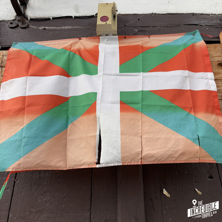 A colorful flag featuring a geometric design with a white cross overlaid on orange, green, and blue triangular sections hangs on a wall. The flag is slightly worn and has a small tear in the middle.