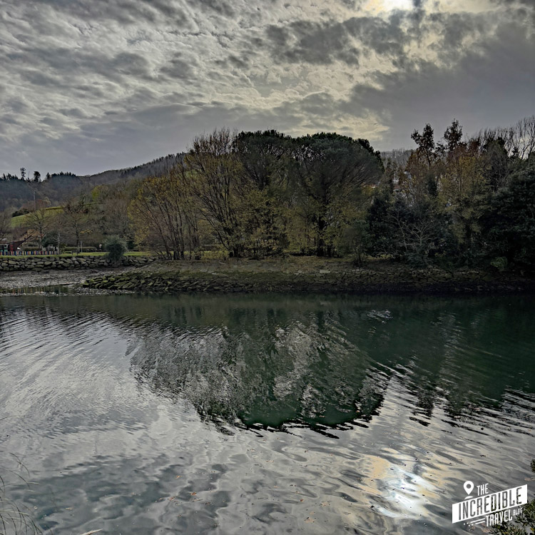 A calm river reflects the cloudy sky and surrounding trees on its surface. The landscape has a serene, overcast atmosphere. A small logo in the corner reads The Incredible Travel.