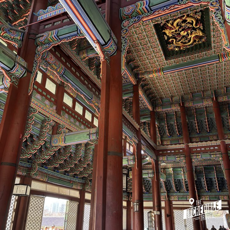 Traditioneller ostasiatischer Tempel mit aufwendig verziertem Holzdach, roten Säulen und farbenfrohen Deckenmalereien in Gold und Grün.