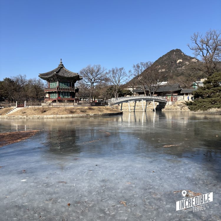 Gefrorener See mit traditionellem asiatischem Pavillon, Steinbrücke und Bergen im Hintergrund bei klarem Himmel