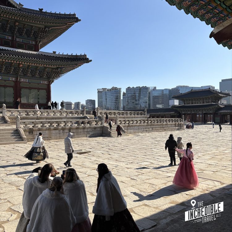 Traditionelle asiatische Tempelanlage mit aufgestuften Dächern, Steinbalustrade und modernen Hochhäusern im Hintergrund. Besucher in traditioneller Kleidung auf dem Platz.