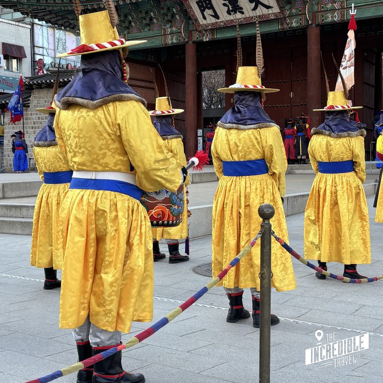 Drei Personen in gelben Gewändern mit blauen Bändern und roten Hüten bei einer traditionellen Zeremonie in einem asiatischen