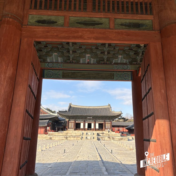 Blick durch rotes Holztor auf traditionelles asiatisches Gebäude mit geschwungenenem Dach in Innenhofanlage.
