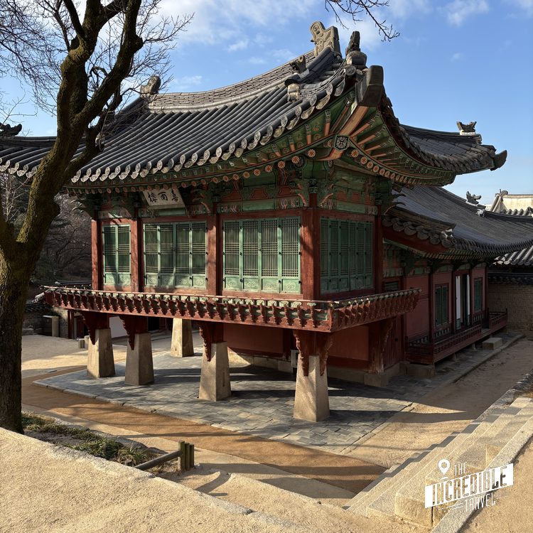 Traditionelles ostasiatisches Gebäude mit geschwungenem Dach, roten Wänden und grünen Türen, umgeben von Bäumen und Steinplatz.