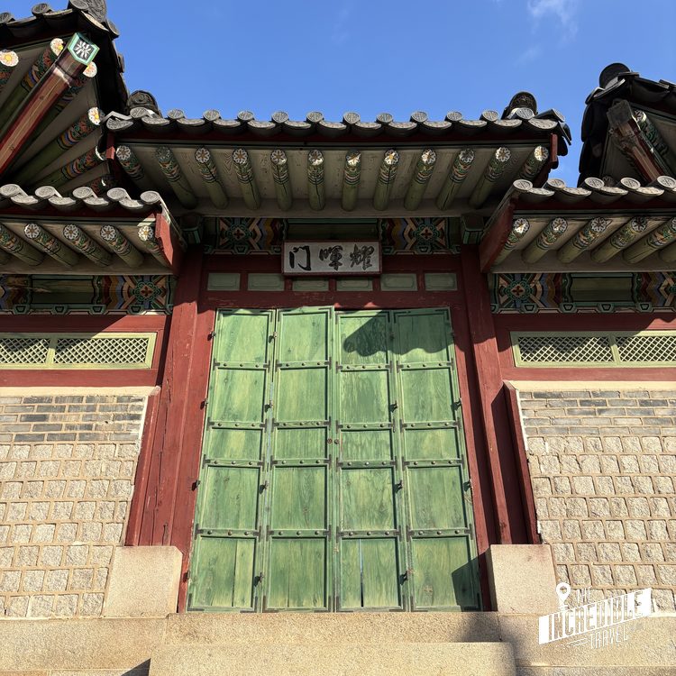 Traditionelles ostasiatisches Gebäude mit grüner Holztür, verziertem Dach und roten Holzelementen unter blauem Himmel.