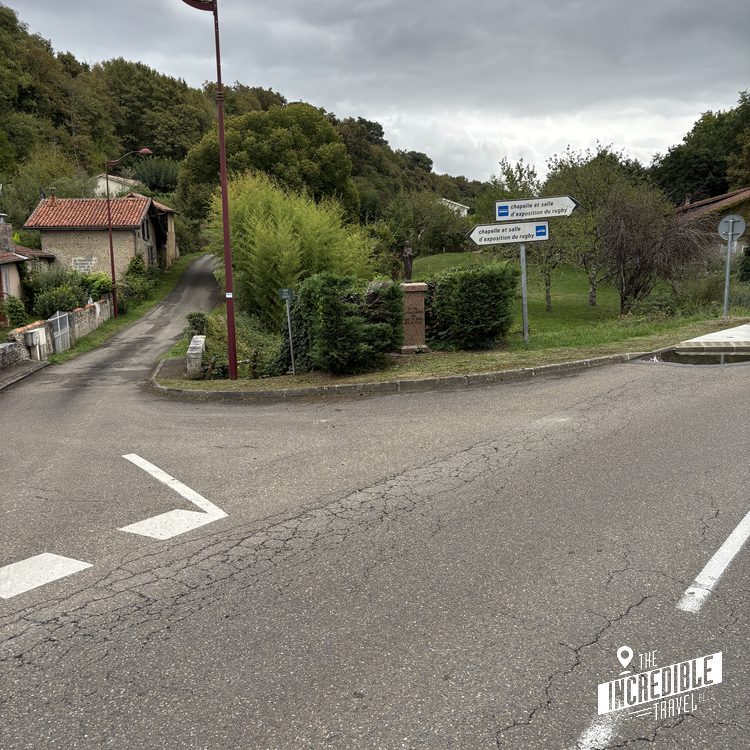 Straßenkreuzung in einem ländlichen Dorf mit Wegweisern, umgeben von grünen Bäumen und traditionellen Steinhäusern.