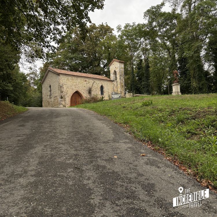 Kleine Steinkirche mit Satteldach und Glockenturm, umgeben von Bäumen und Grünflächen, Zufahrtsweg im Vordergrund