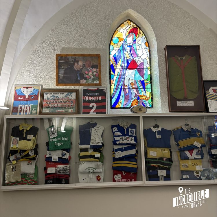 Ausstellungsregal mit historischen Rugby-Trikots und Ausrüstung, darüber Buntglasfenster und Fotos an weißer Wand
