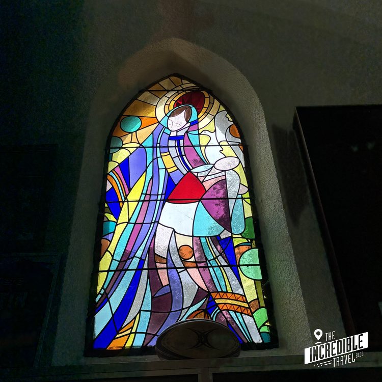 Buntes modernes Buntglasfenster mit abstrakten geometrischen Formen und einer Marienfigur mit Heiligenschein in einer Kirchennische.