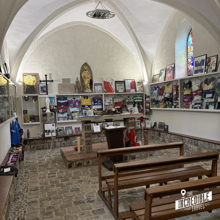 Kapelleninneres mit Holzbänken, weißen Gewölbedächern, religiösen Kunstwerken an den Wänden und Erinnerungsstücken in Vitrinen.