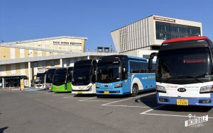 Parkplatz mit mehreren touristischen Bussen in verschiedenen Farben vor einem Gebäude mit koreanischer Beschriftung unter blauem Himmel.