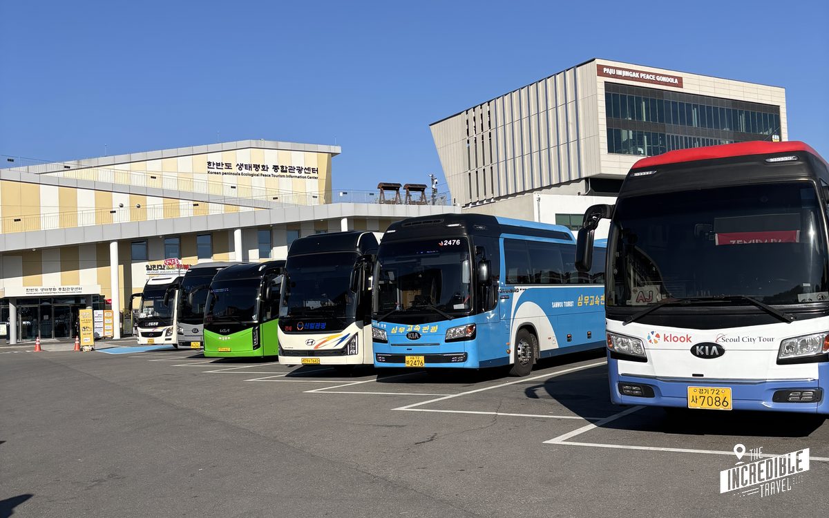 Parkplatz mit mehreren touristischen Bussen in verschiedenen Farben vor einem Gebäude mit koreanischer Beschriftung unter blauem Himmel.