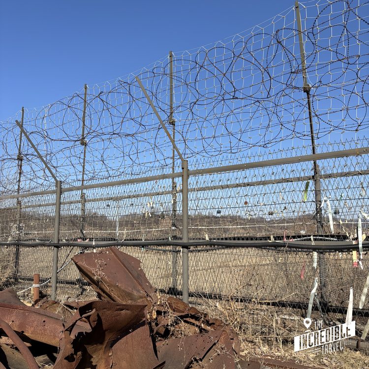 Rostiger Metallschrott vor hohem Metallzaun mit Stacheldraht unter blauem Himmel
