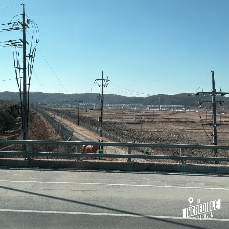 Blick über eine Brücke mit Metallgeländer auf ausgedehnte Weideflächen, Strommasten und bewaldete Hügel unter blauem Himmel.