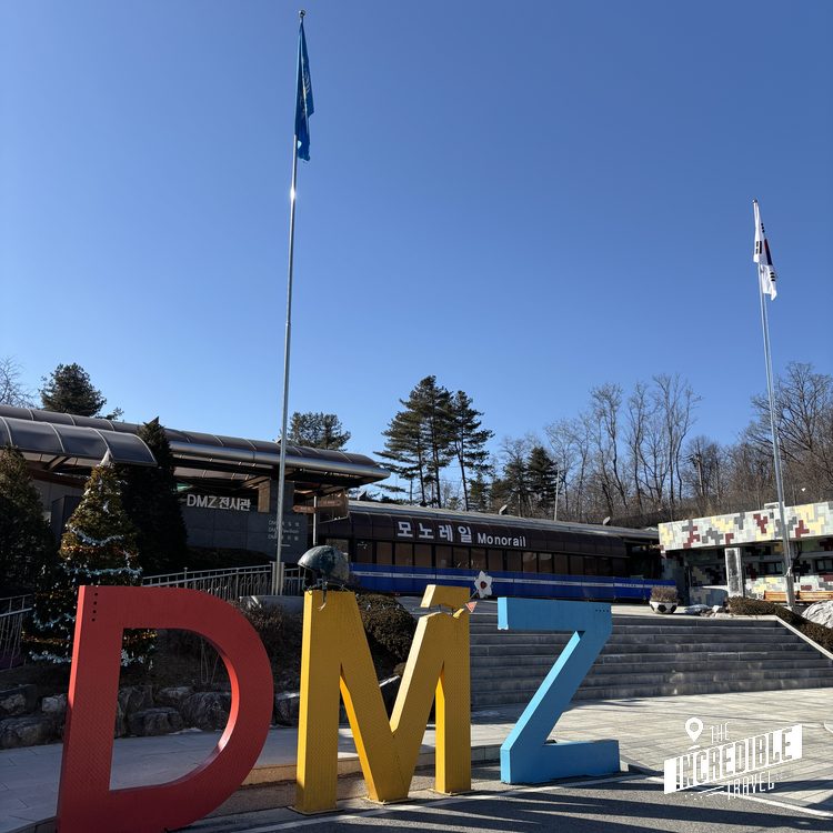 Bunte Großbuchstaben DMZ vor einer Monorail-Station mit Flaggen und Bäumen unter blauem Himmel