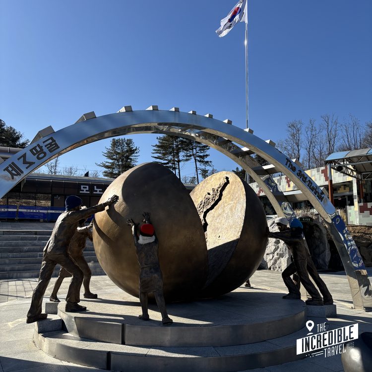 Bronzeskulptur von drei Figuren, die einen großen Stein tragen, unter blauem Bogen, koreanische Flagge im Hintergrund.