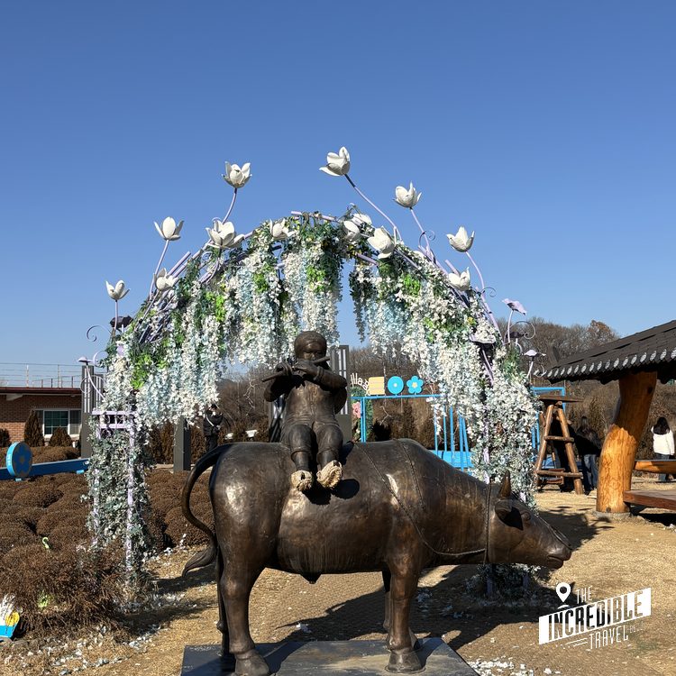 Bronzeskulptur eines Kindes auf einem Rind unter einer blütengeschmückten Laube aus weißen Blüten.