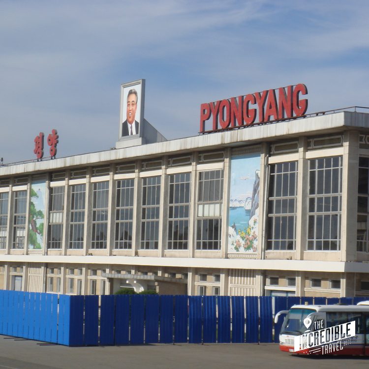 Großes Gebäude mit "Pyongyang"-Schriftzug und Portrait an der Fassade, dahinter blauer Himmel