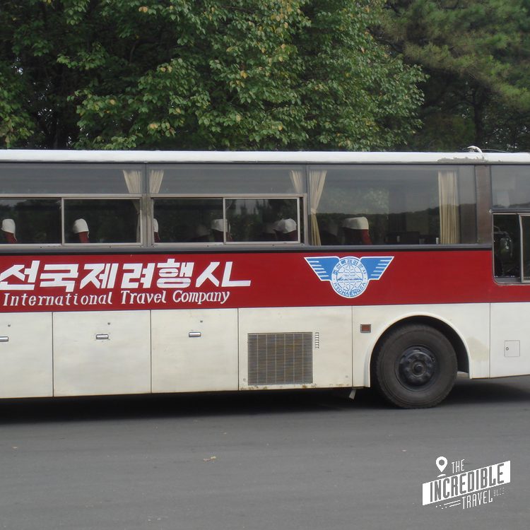Weißer und roter Reisebus mit koreanischen Schriftzeichen und englischer Aufschrift "International Travel Company" vor grünem Laubwald.