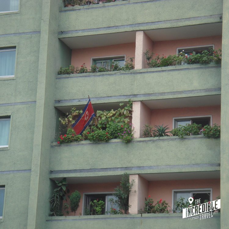 Mehrgeschossiges Wohngebäude mit grüner Fassade, rosa Balkonen, Blumenkästen und einer Flagge an einem Balkon.