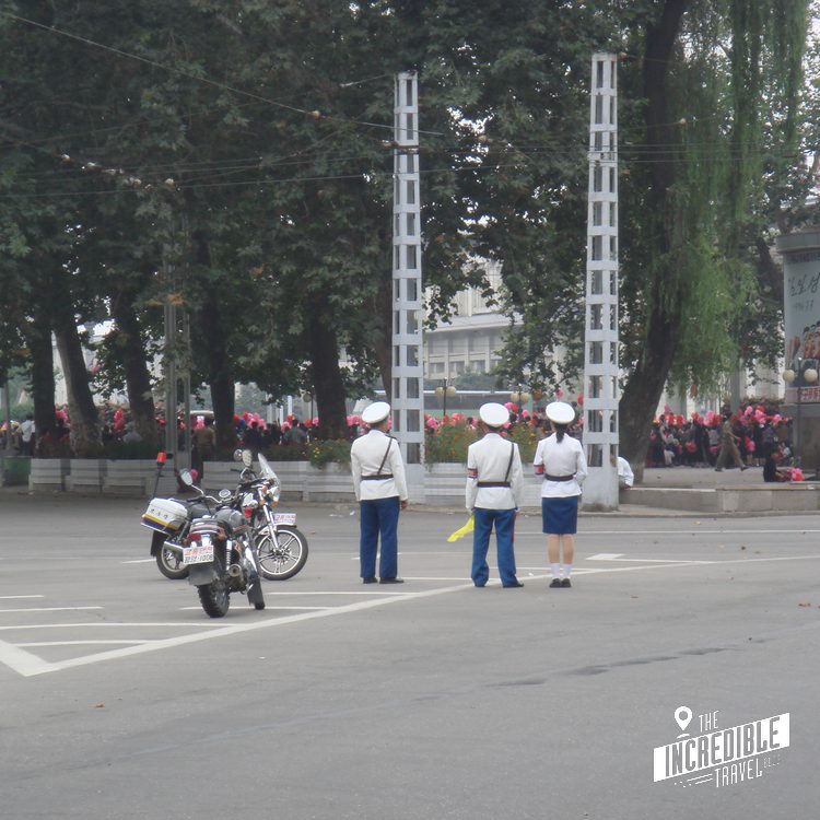 Militärparade mit Motorrad und uniformierten Soldaten auf breitem Platz, umgeben von Bäumen und Zuschauern.