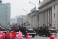 Militärparade mit Lastwagen und Raketenwerfern auf breiter Straße, rote Blumen im Vordergrund, historische Gebäude und Flaggen