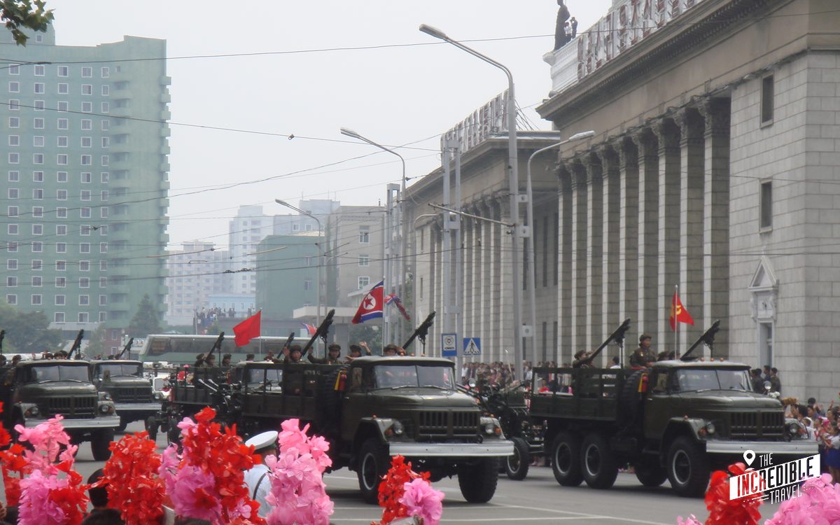 Militärparade mit Lastwagen und Raketenwerfern auf breiter Straße, rote Blumen im Vordergrund, historische Gebäude und Flaggen