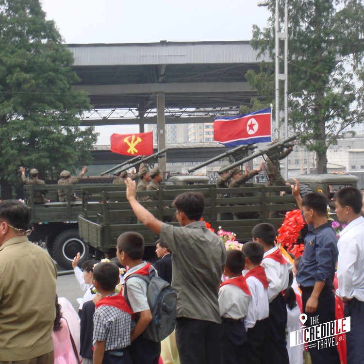 Militärparade mit Truck, Flaggen und jubelnder Menschenmenge unter einer Brücke
