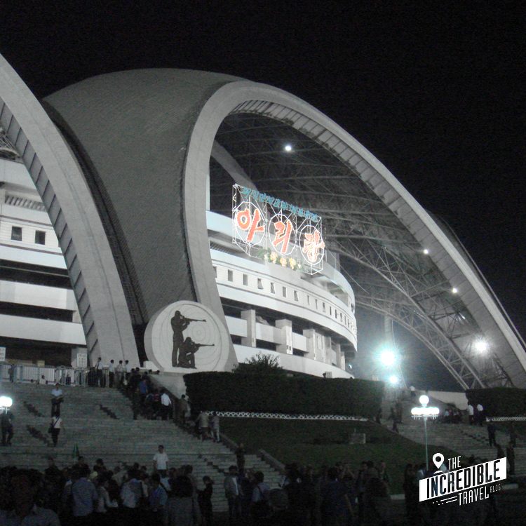 Stadion bei Nacht mit geschwungener Architektur, beleuchtet von Flutlicht, Besucher auf Treppen