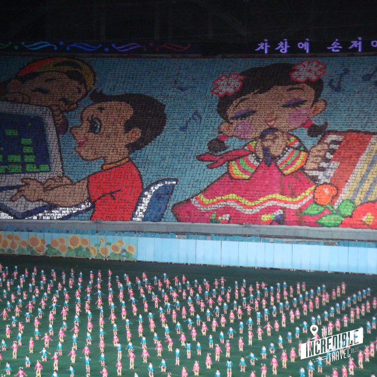 Großes Mosaikbild mit koreanischen Schriftzeichen zeigt Personen in farbigen Gewändern. Darunter Tausende kleine rosa Figuren in Formation auf grünem Untergrund.