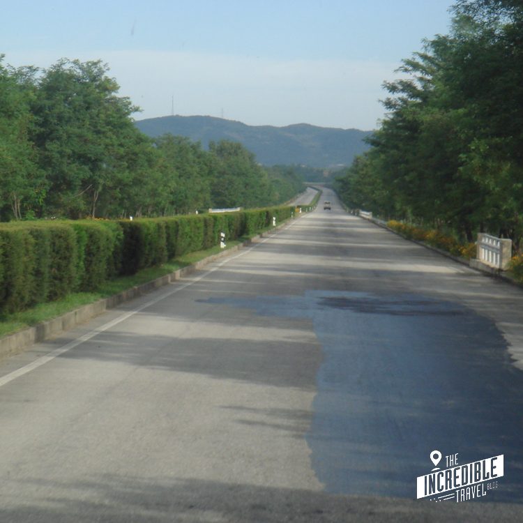 Gerade Landstraße zwischen grünen Hecken mit Bergen im Hintergrund und Pfütze auf der Fahrbahn.