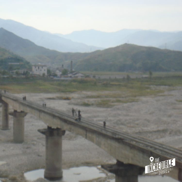 Betonbrücke mit Pfeilern über breitem Fluss in Berglandschaft mit Fabrik und Siedlung im Hintergrund