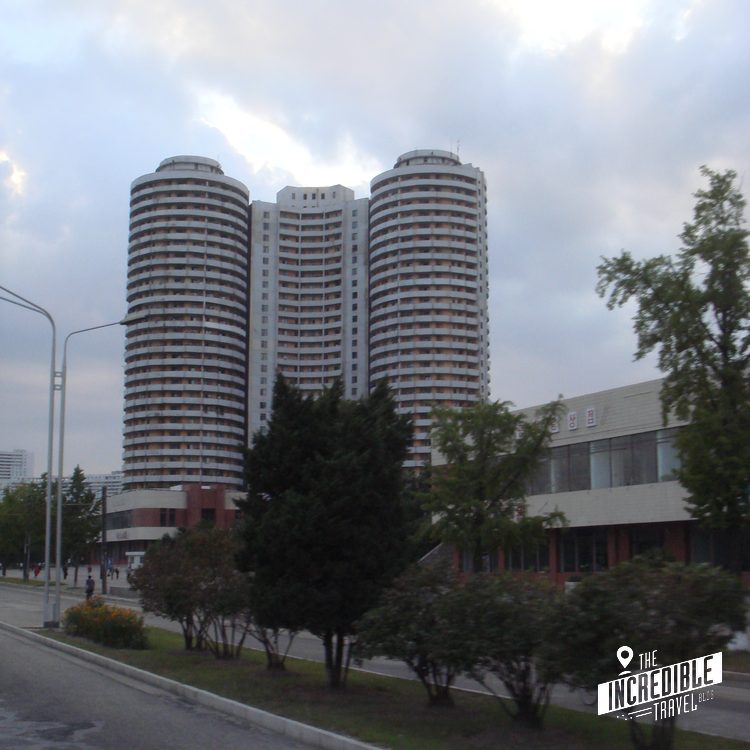 Drei moderne Hochhäuser mit gebogenen Fassaden neben einem mehrstöckigen Gebäude, umgeben von Grünflächen und Straßenlaternen unter bewölktem Himmel.