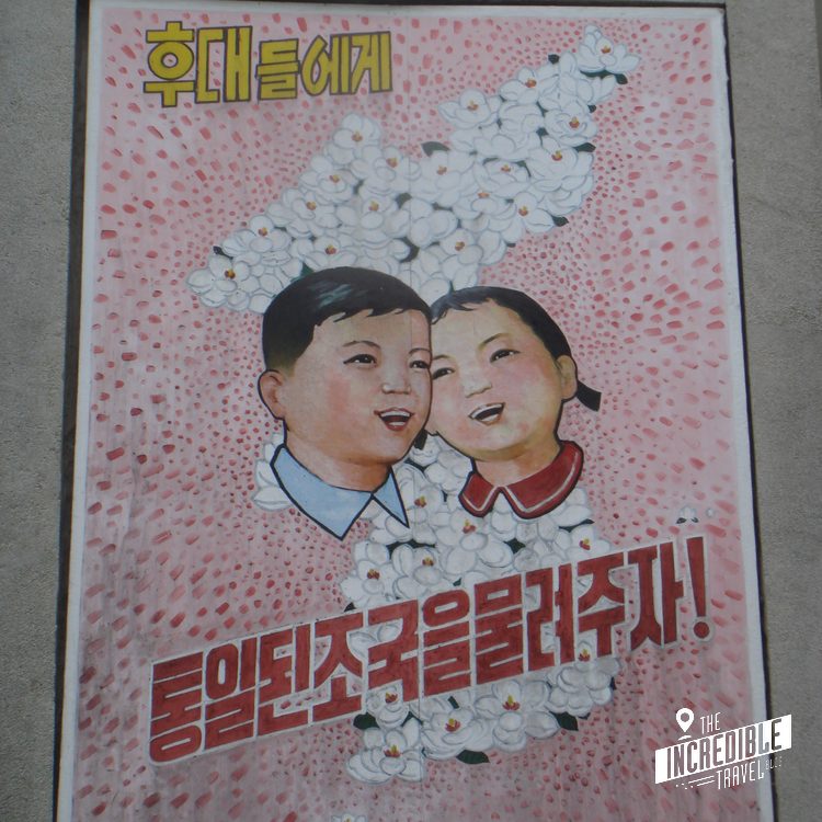 Propagandaplakat mit koreanischem Text, zwei lächelnden Gesichtern und dekorativem weißen Muster auf rosa Hintergrund.