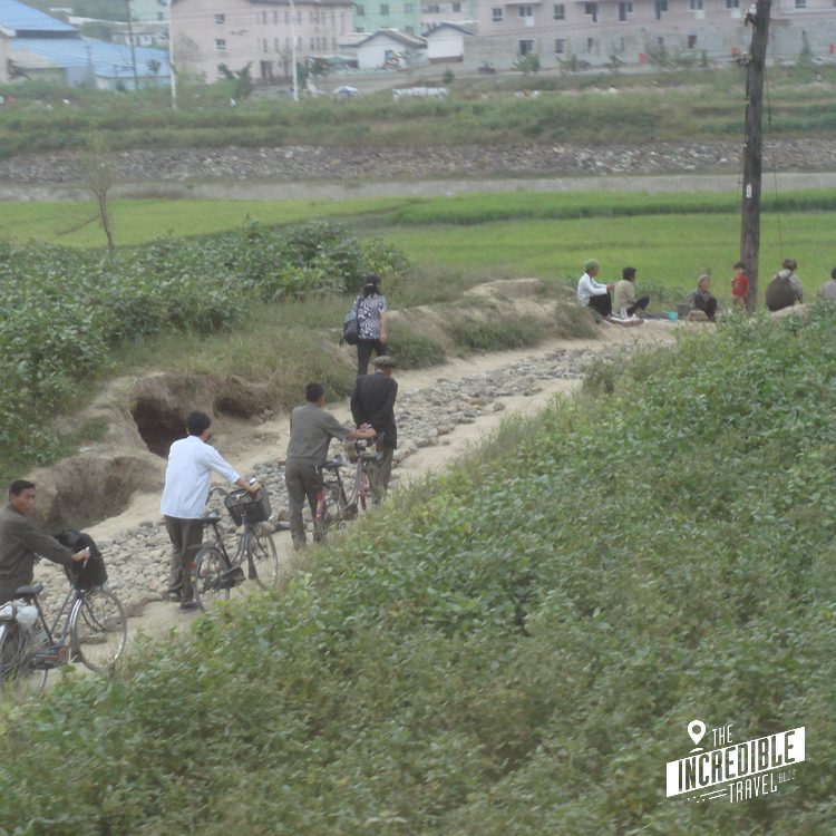 Gruppe von Menschen mit Fahrrädern auf einem Feldweg zwischen grünen Feldern und Wohnblöcken im Hintergrund.